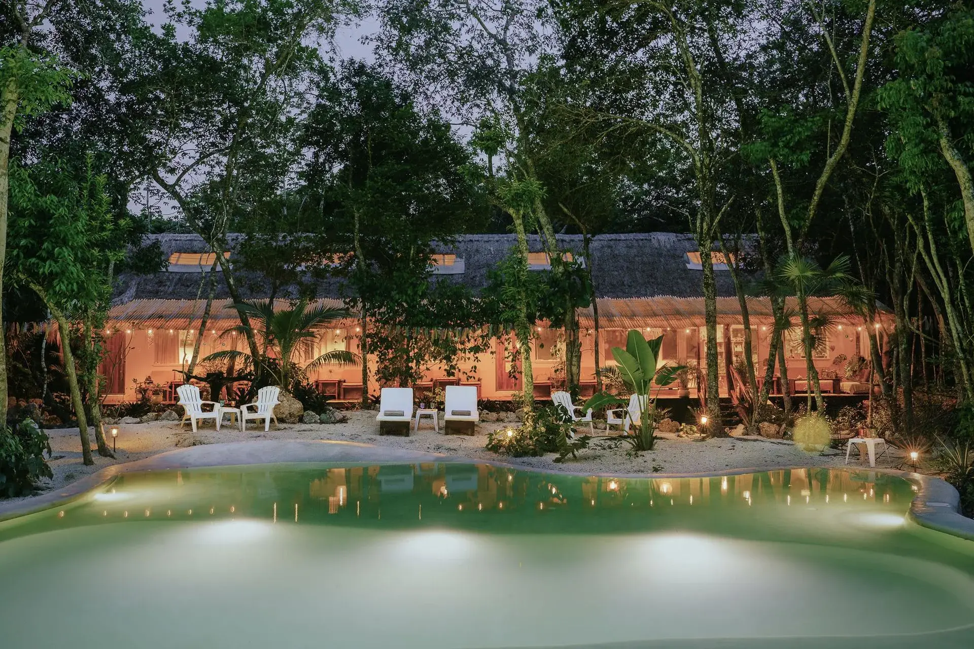 The main veranda at Casa Arkaana at dusk — warm interior light, cenote-fed pool in the foreground, jungle canopy behind